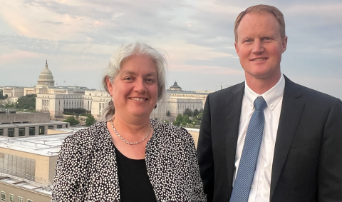 Joan Ramage and Jed Hancock pose for a photo on a rooftop in Washington, D.C. Behind them is the Capital building and the sun is setting.